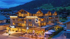A multi-story hotel with a stone facade, outdoor seating, and a mountain backdrop. - MOUNTAIN LUIS - HOTEL | SUITES | PENTHOUSES (Kaprun)