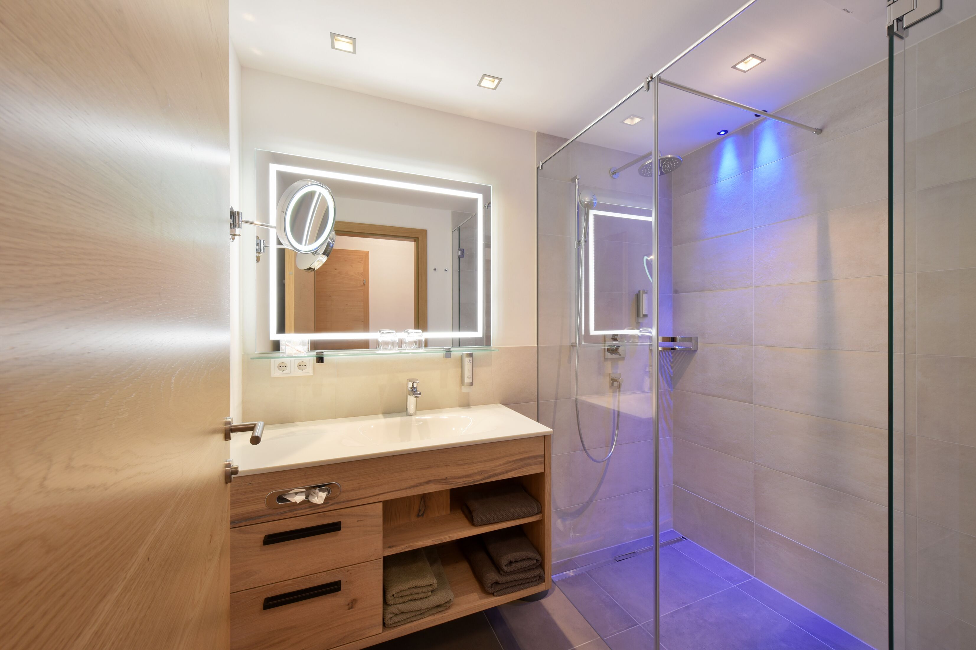 A modern bathroom with a wooden vanity, a glass shower enclosure, and a mirror with built-in lighting.