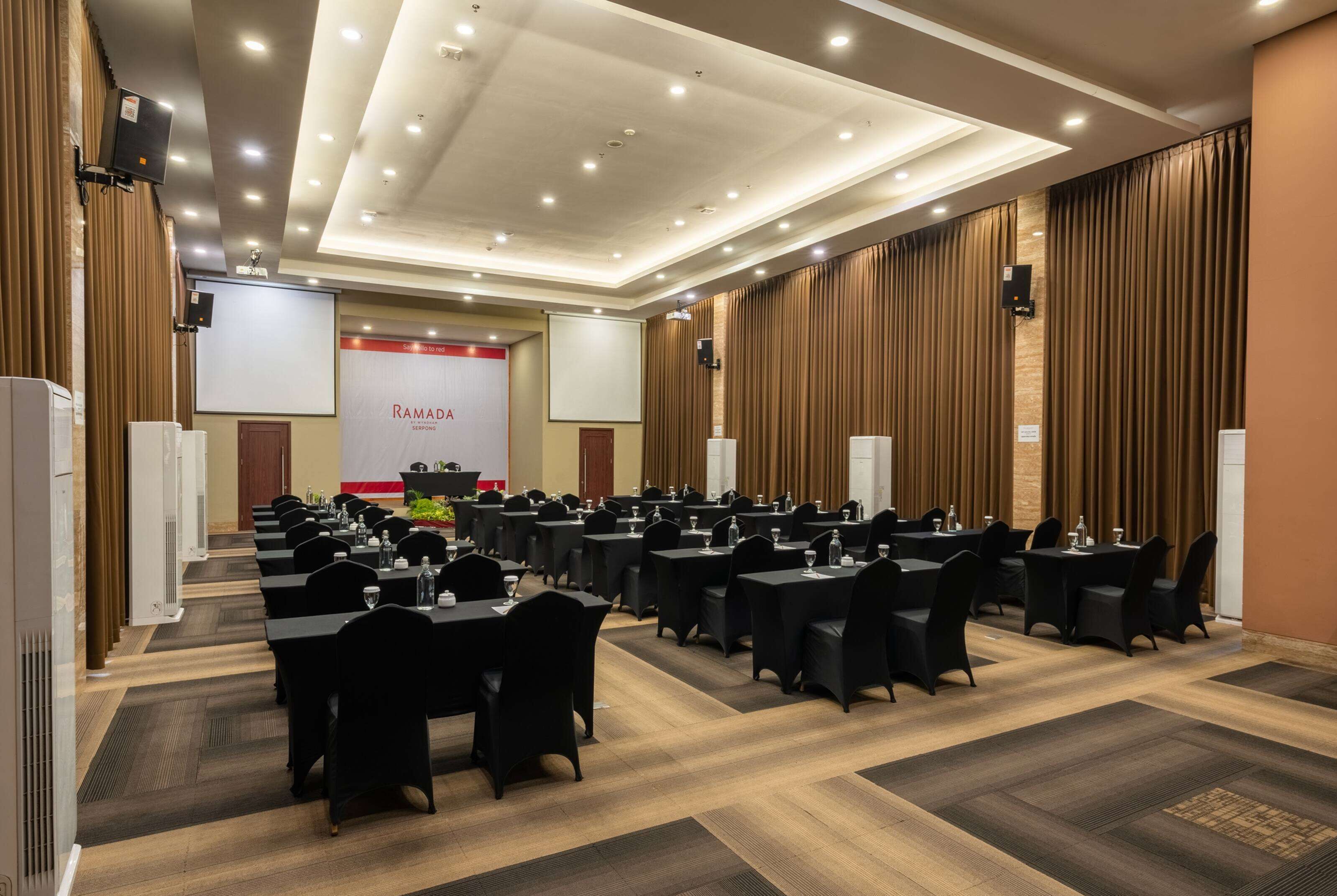 A conference room with multiple tables and chairs, a podium, and a screen displaying "RAMADA".