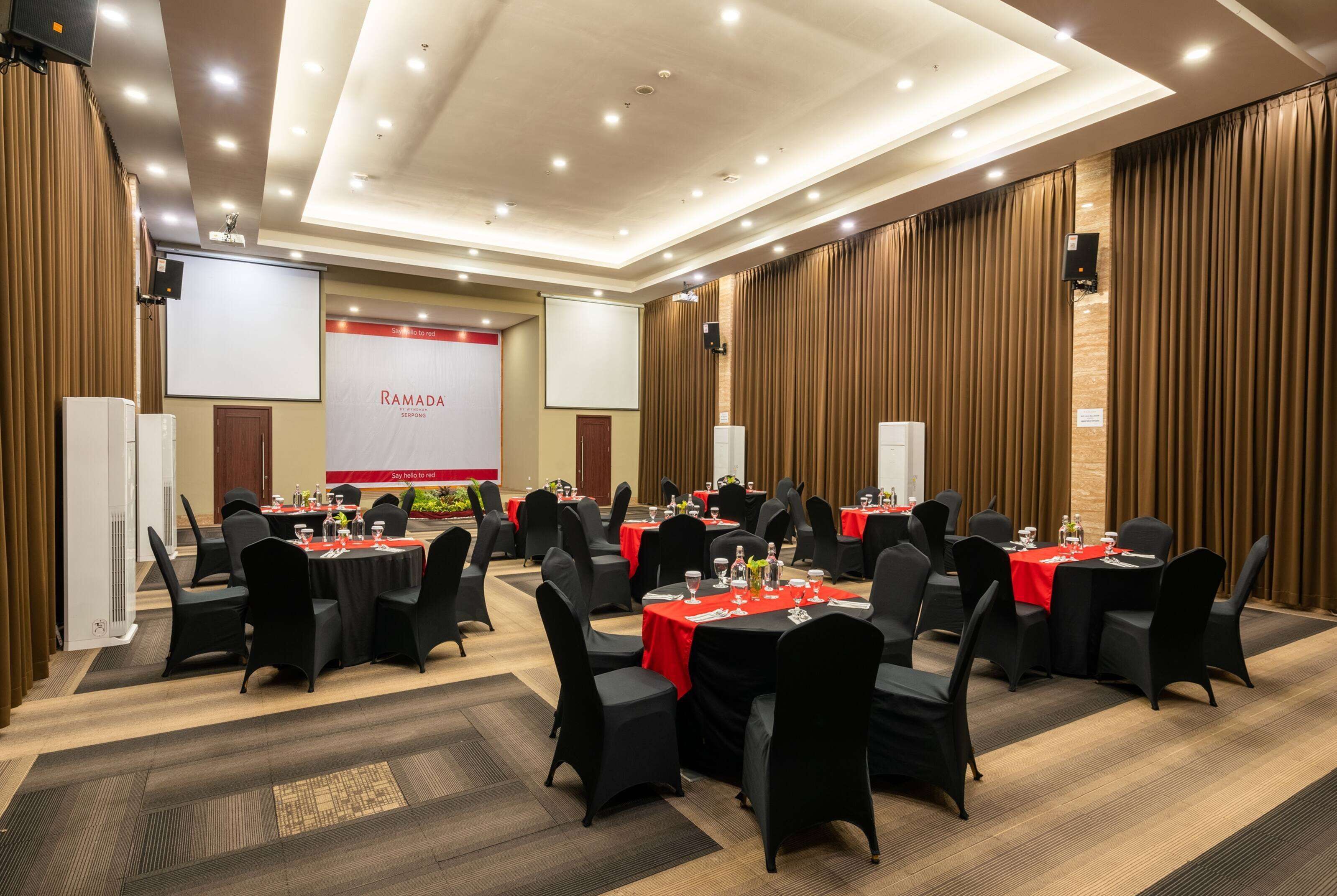 A conference room with multiple tables, black chairs, and red tablecloths. There are screens and speakers on the walls.