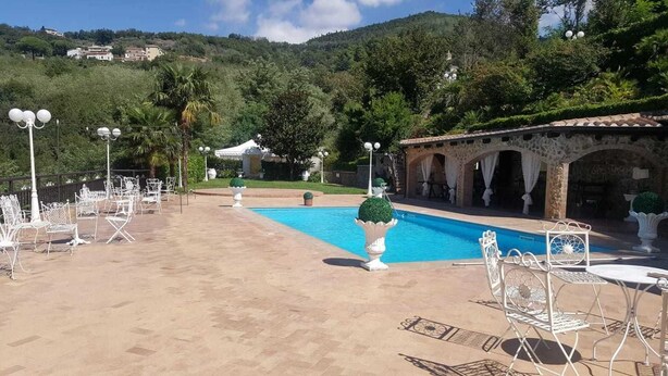 A pool area with white metal chairs and tables, a stone bridge, and a view of a hill with buildings in the background. - B&B La Mimosa (Cava de' Tirreni)