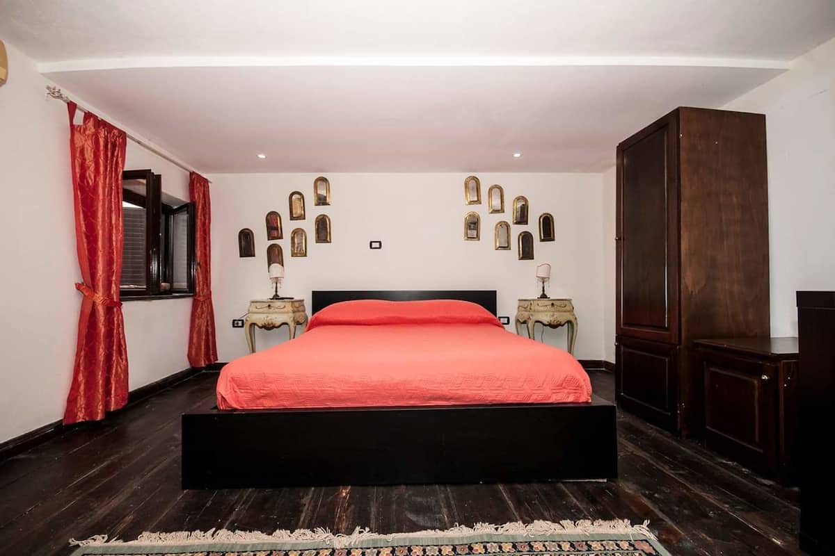 Een slaapkamer met een groot bed, een donkere houten vloer, rode gordijnen en een wand versierd met lijstwerken.