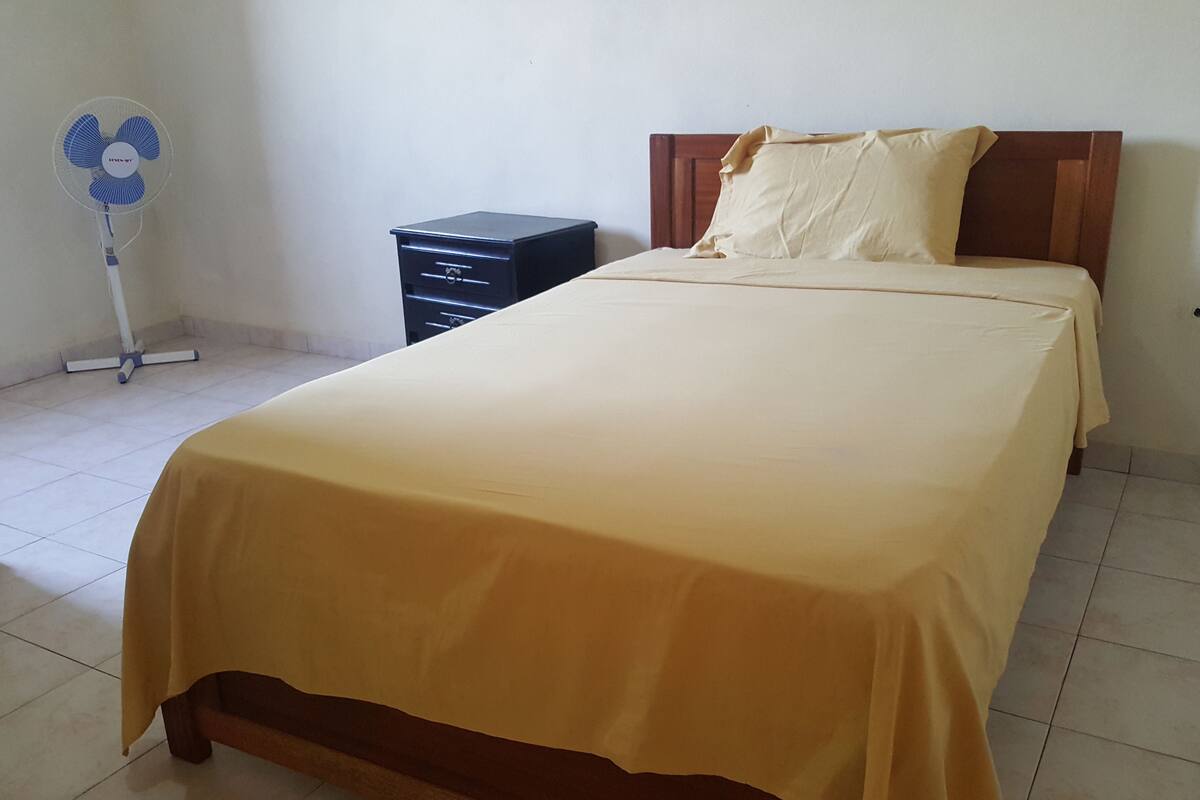 Quarto com cama, cabeceira de madeira, mesa de cabeceira e ventilador.