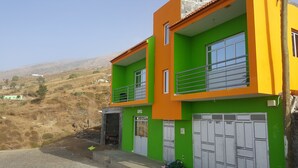 A two-story building with a green and orange facade, a balcony, and a garage with three doors labeled "Casa," "Monte," and "Branco." - Casa Tchichi (São Filipe)