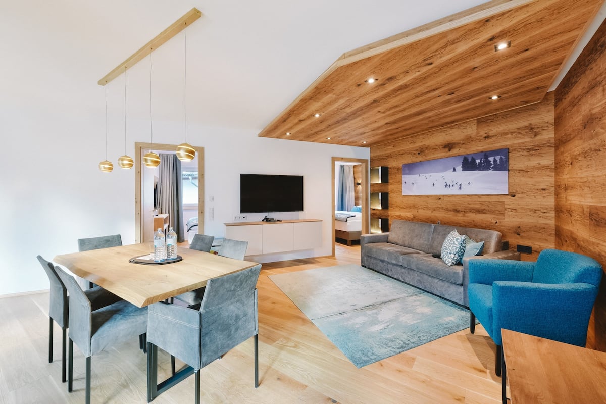 A modern living room with a wooden ceiling, a flat-screen TV, a grey sofa, a blue armchair, and a dining area with a wooden table and chairs.
