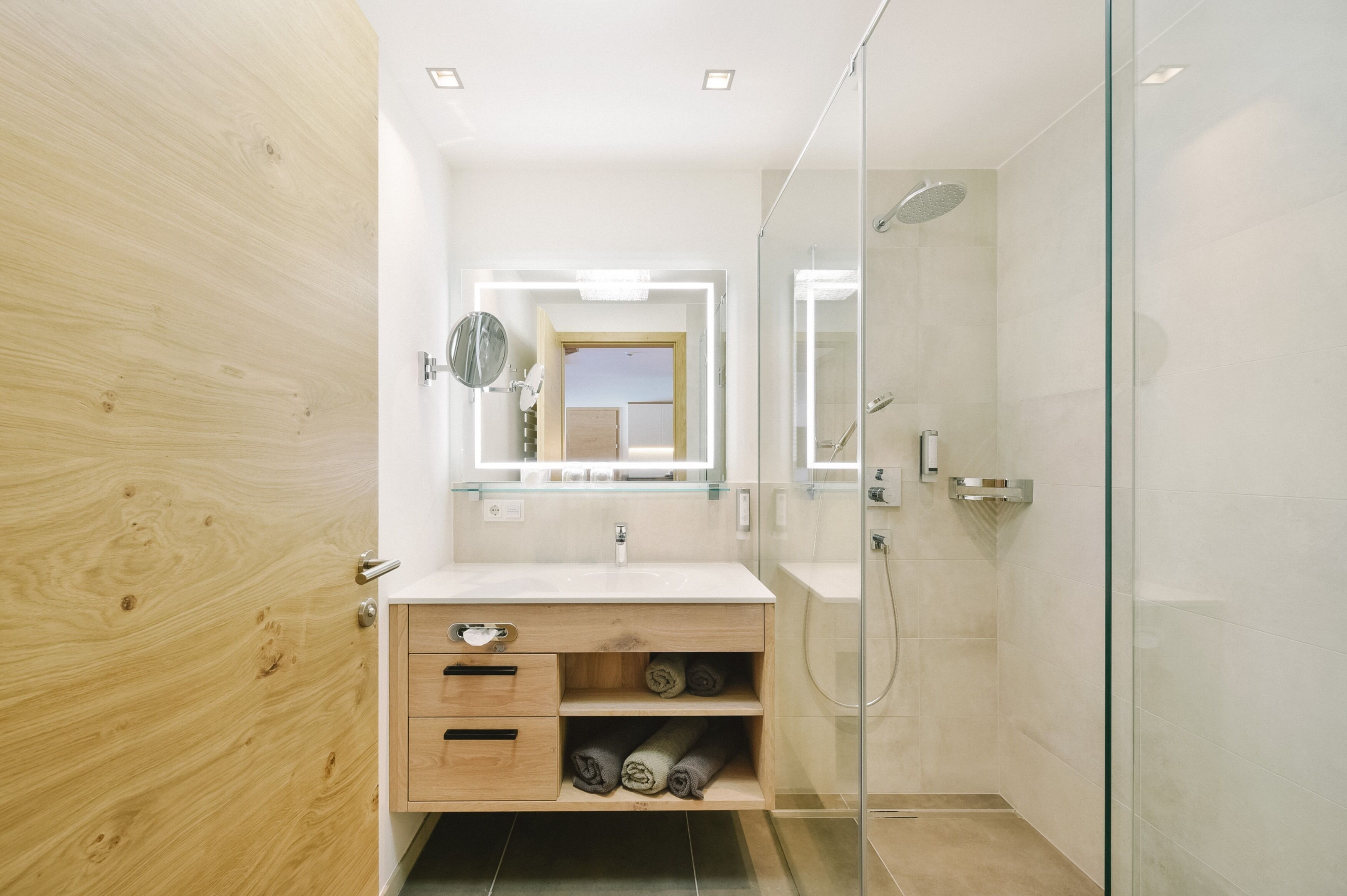 A modern bathroom with a wooden vanity, a glass shower enclosure, and a mirror with integrated lighting.