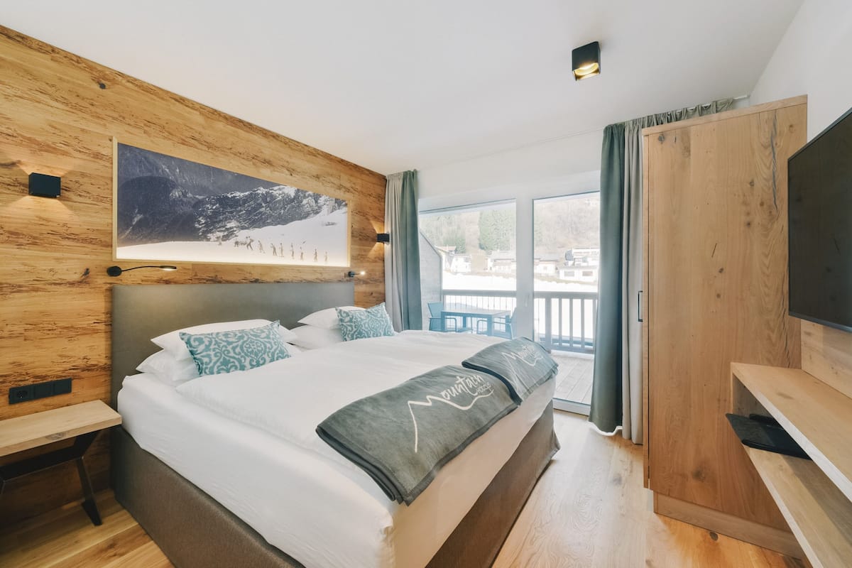 A modern hotel room with a large bed, wooden headboard, a mounted TV, and a balcony with a view of the town.