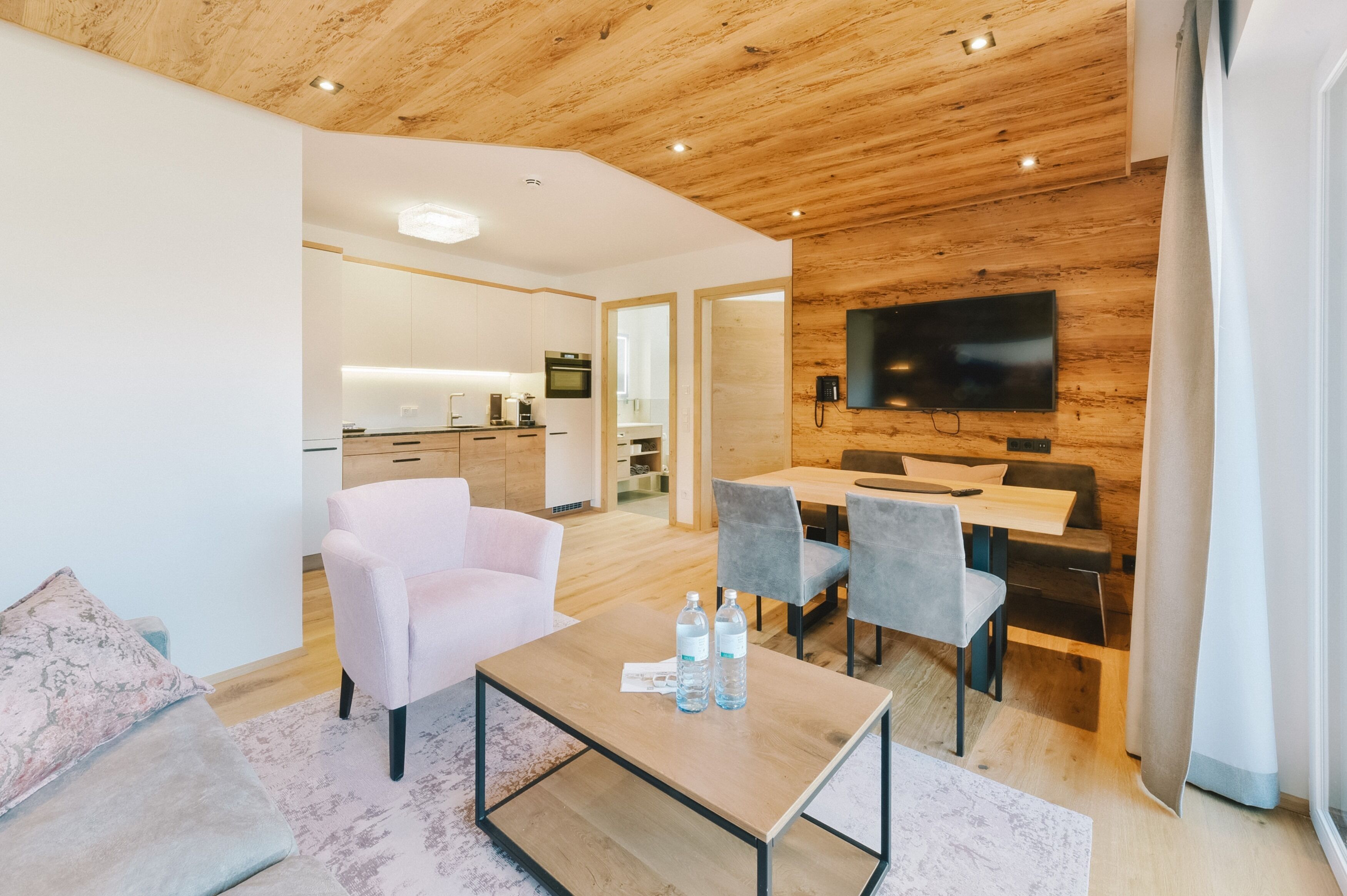 A modern living room with a wooden ceiling, a flat-screen TV mounted on the wall, a dining area with grey chairs, and a kitchen in the background.