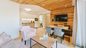 A modern living room with a wooden ceiling, a flat-screen TV mounted on the wall, a dining area with grey chairs, and a kitchen in the background.