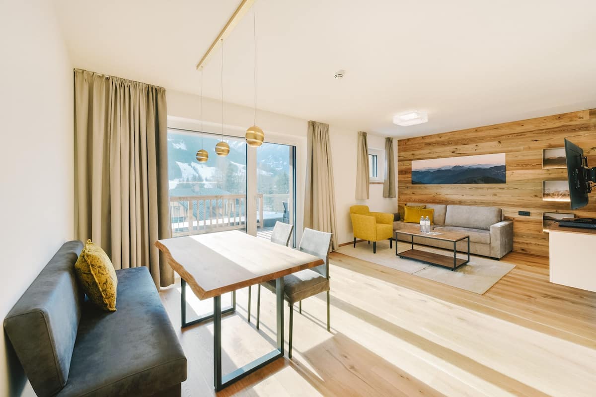 A modern living room with a wooden floor, a sofa, a dining table, and a large window with curtains.
