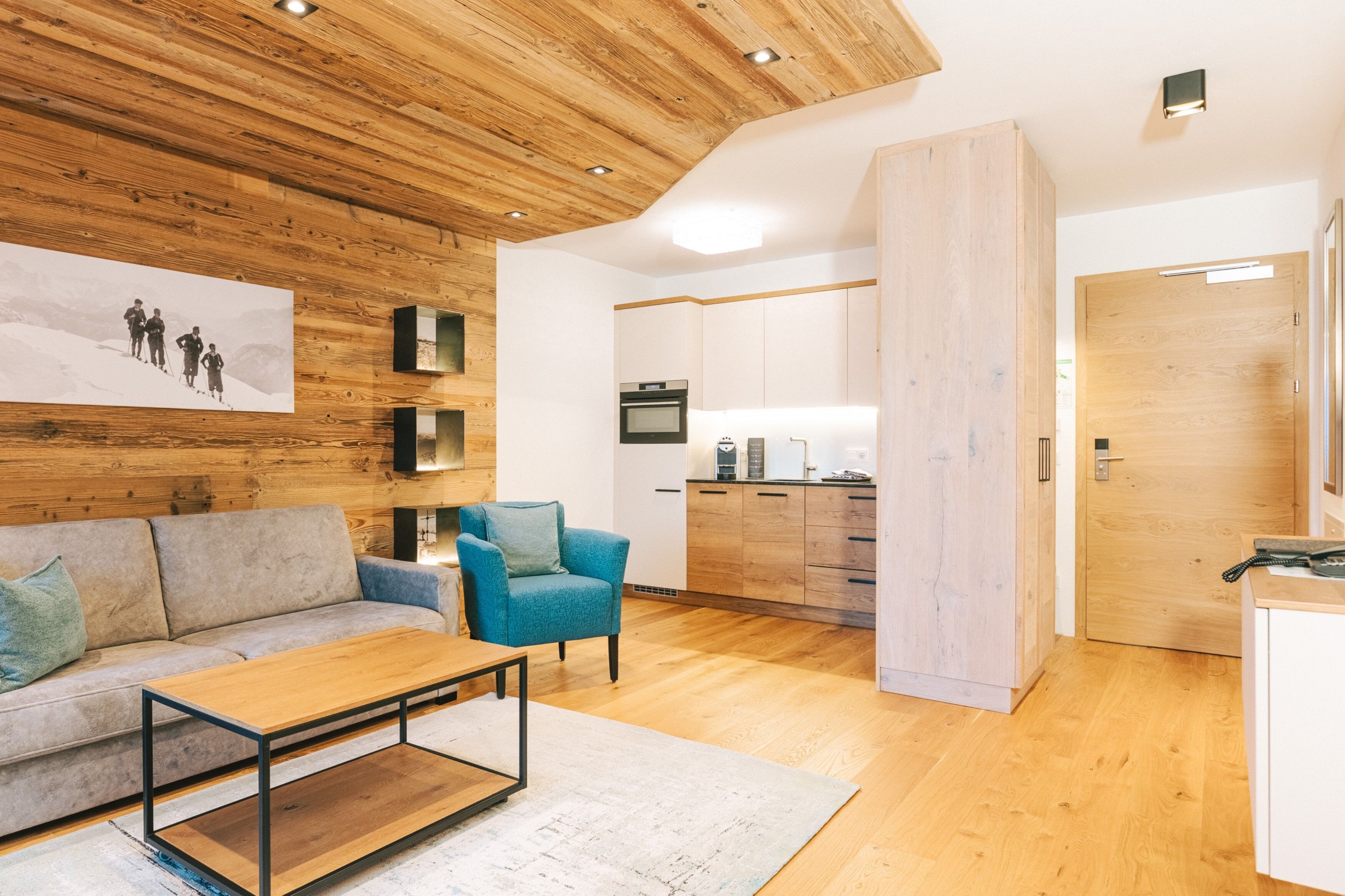 A modern living room with a wooden ceiling, a sofa, a coffee table, and a kitchen area with cabinets and appliances.