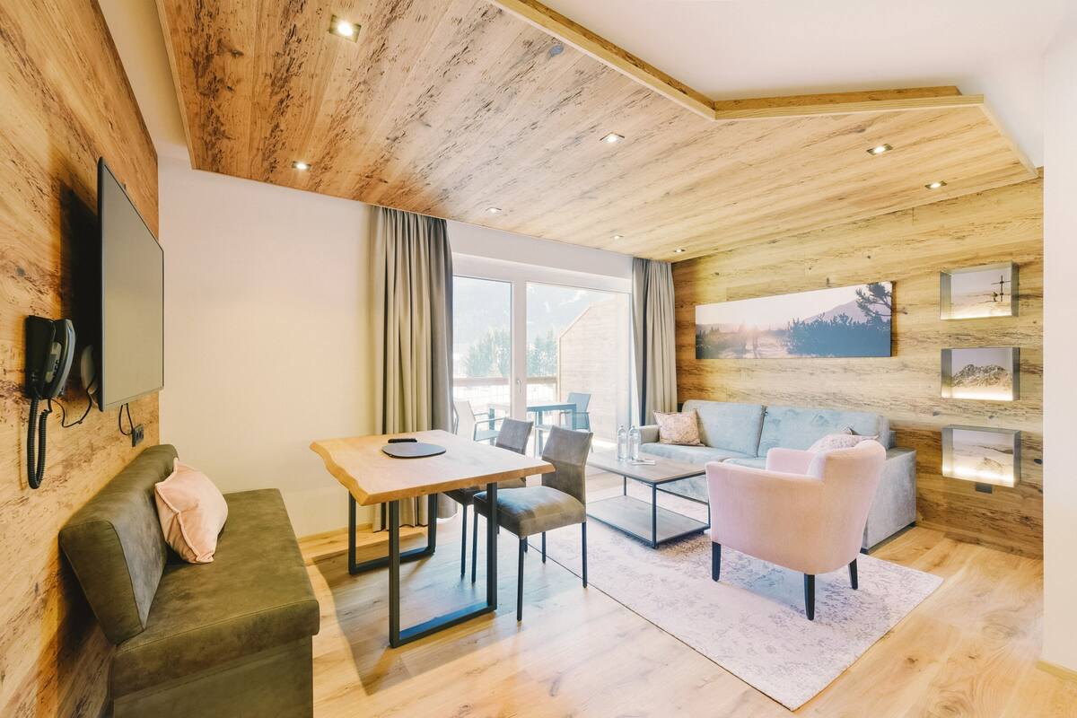 A modern living room with a wooden ceiling, a flat-screen TV mounted on the wall, a dining table with chairs, a sofa, and a coffee table.