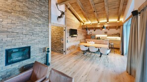 A modern interior with a stone wall, wooden ceiling beams, a dining area with white chairs, and a kitchen with a wooden countertop. - Venediger Lodge (Neukirchen am Grossvenediger)