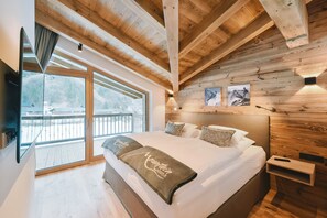 Un dormitorio con una cama grande, vigas de madera en el techo, un televisor montado en la pared y un balcón con vistas a las montañas.