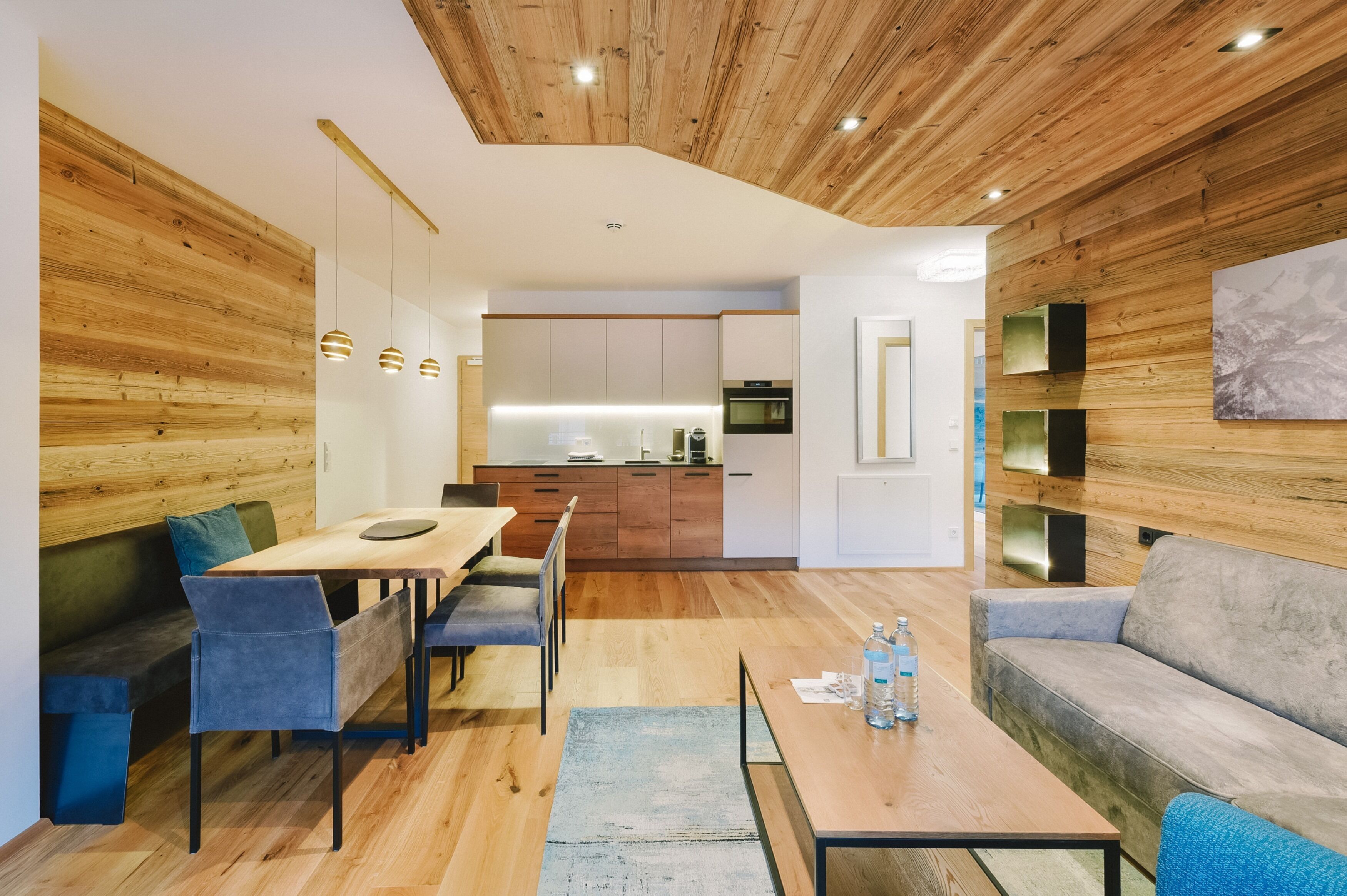A modern living room with a wooden ceiling, a dining area with a table and chairs, and a kitchen in the background.