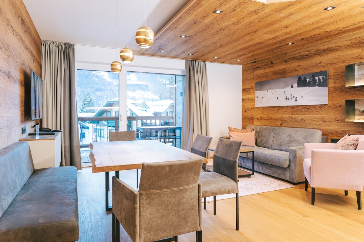 A modern living room with a wooden ceiling, a grey sofa, a dining table with chairs, and a large window with a view of a building and mountains.