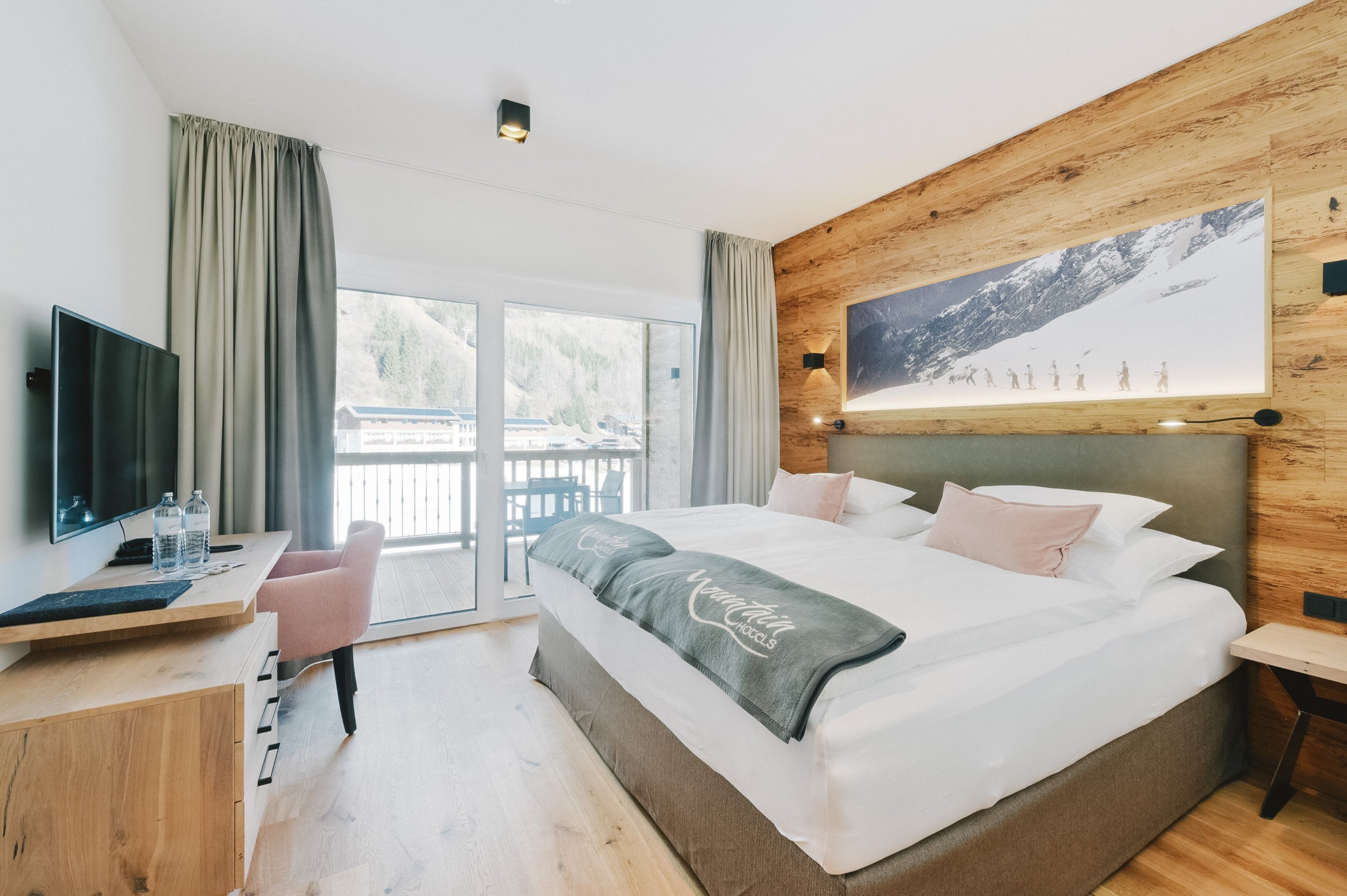 Une chambre d’hôtel moderne dotée d’un grand lit, d’un bureau avec un ordinateur, d’une télévision et d’un balcon offrant une vue sur les montagnes.