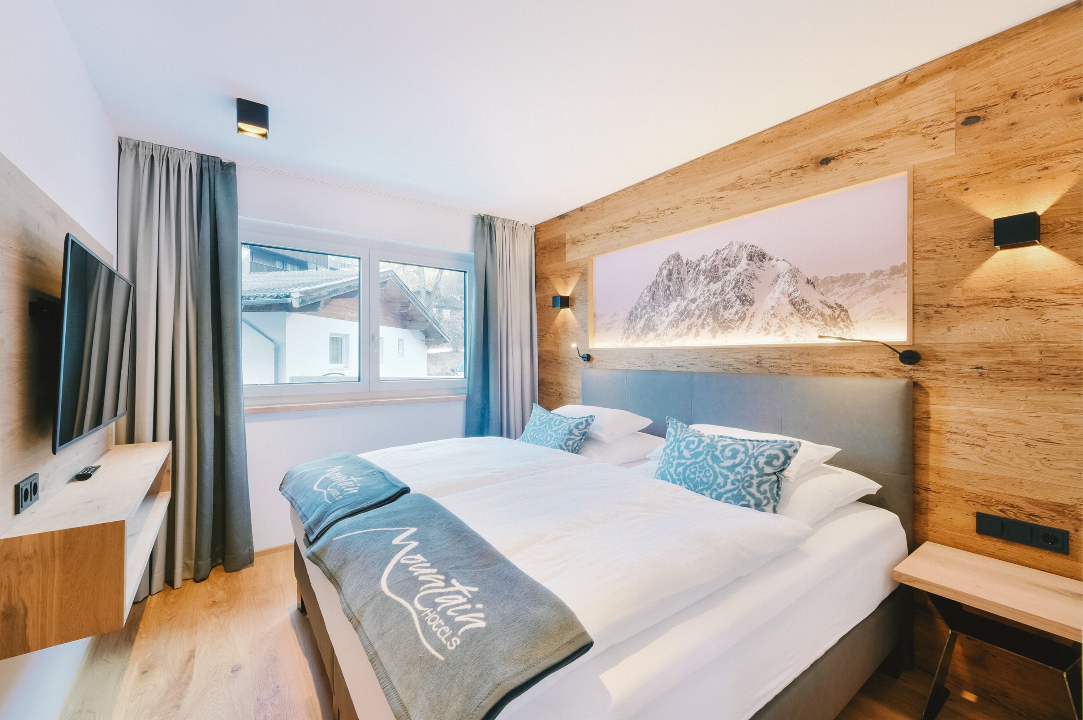A modern hotel room with a large bed, a mounted TV, a window with curtains, and a mountain landscape painting on the wall.
