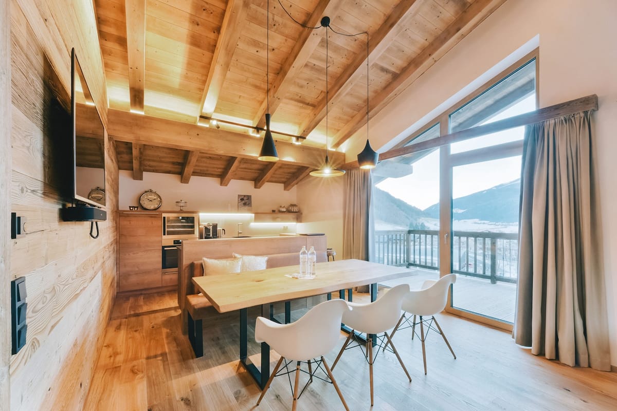A modern kitchen with wooden beams, a dining table with chairs, and a large window with a view of mountains.