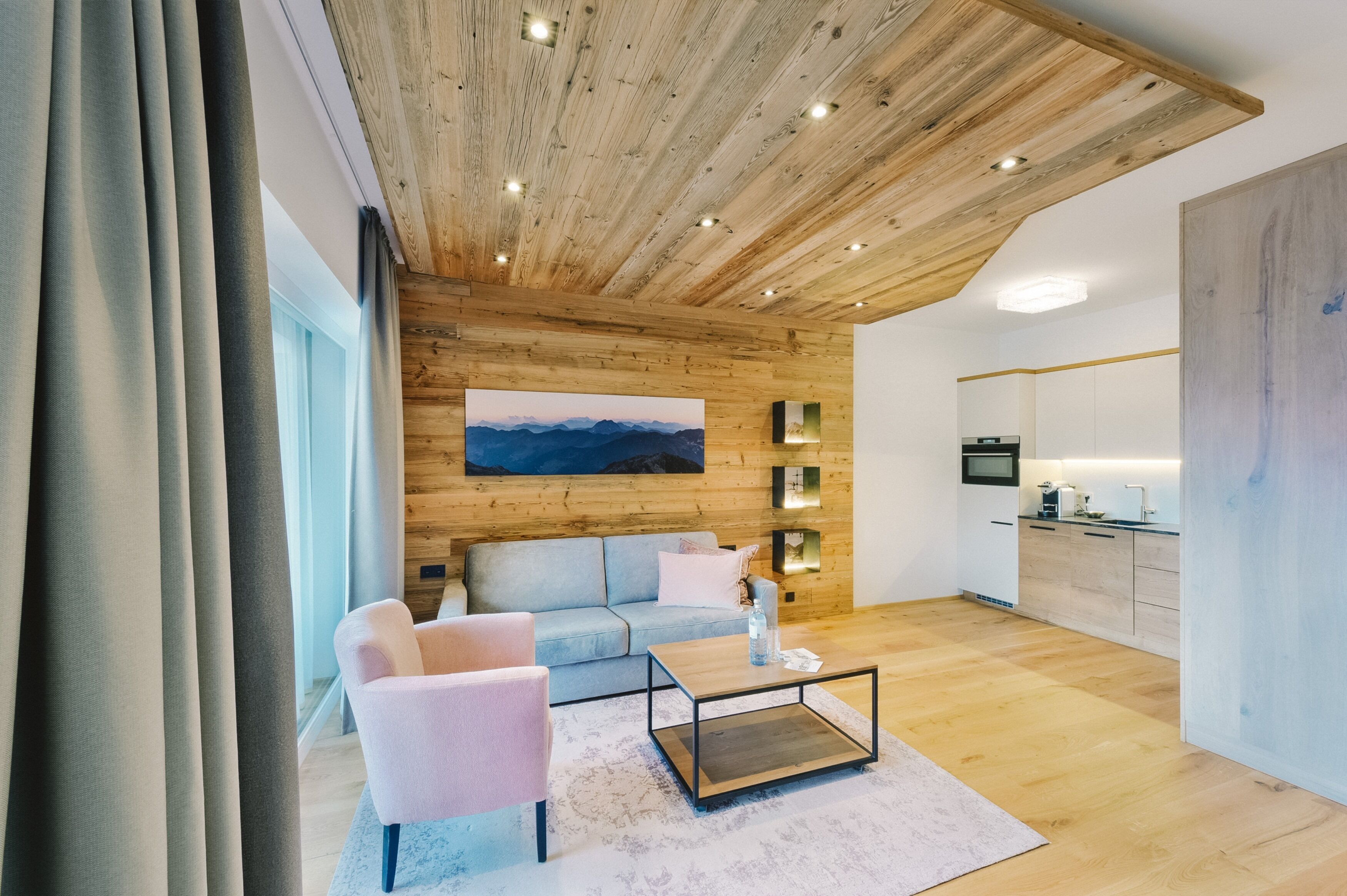 Un salon moderne avec un plafond en bois, un grand tableau, un canapé, une table basse et un coin cuisine doté d’un plan de travail et d’appareils électroménagers.