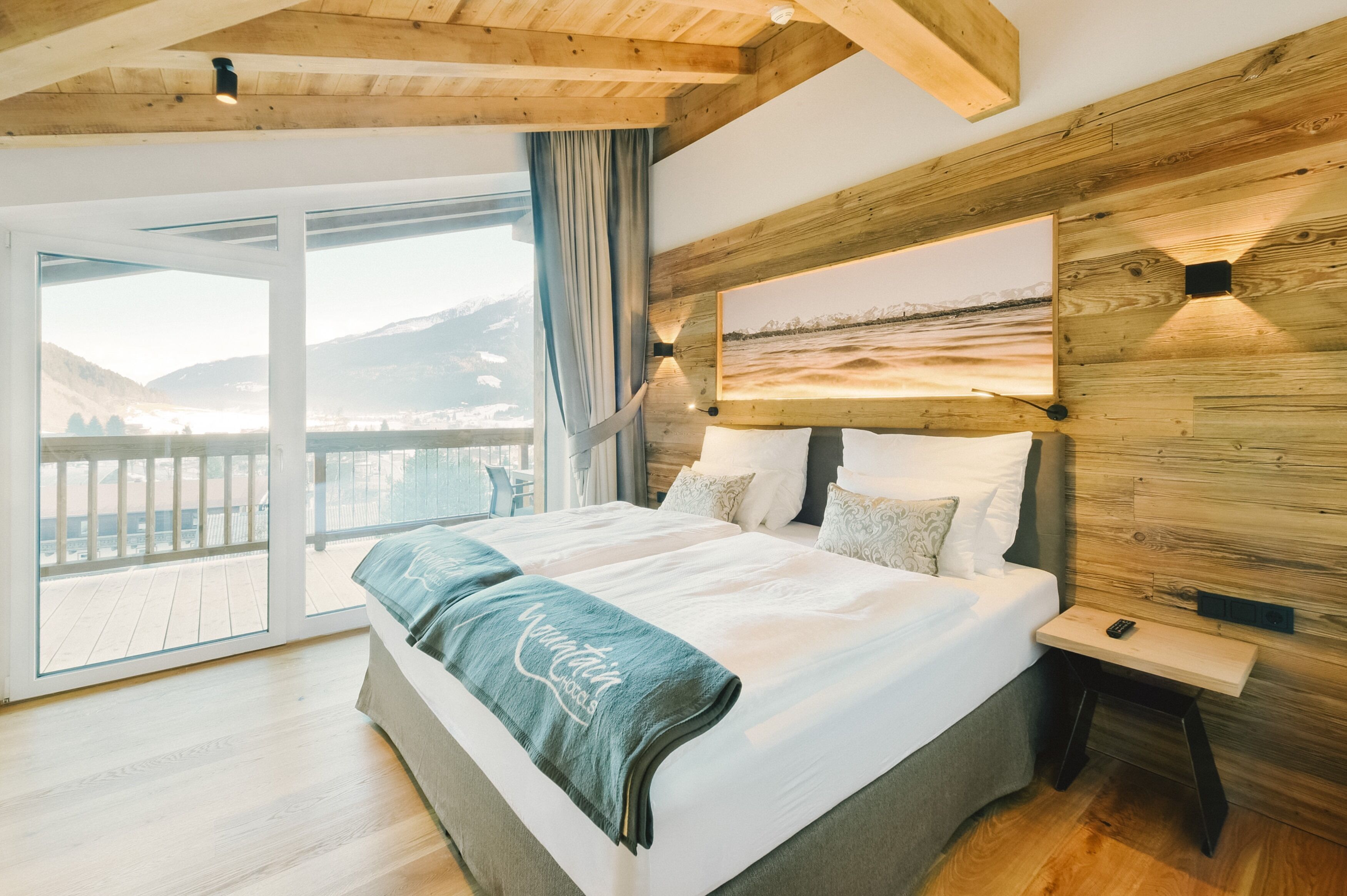 A bedroom with a large bed, a nightstand, a painting on the wall, and a view of the mountains through the window.