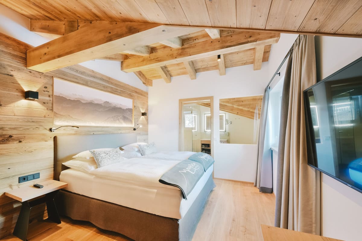 A modern bedroom with a wooden ceiling, a large bed, a bedside table, and a large window with curtains.