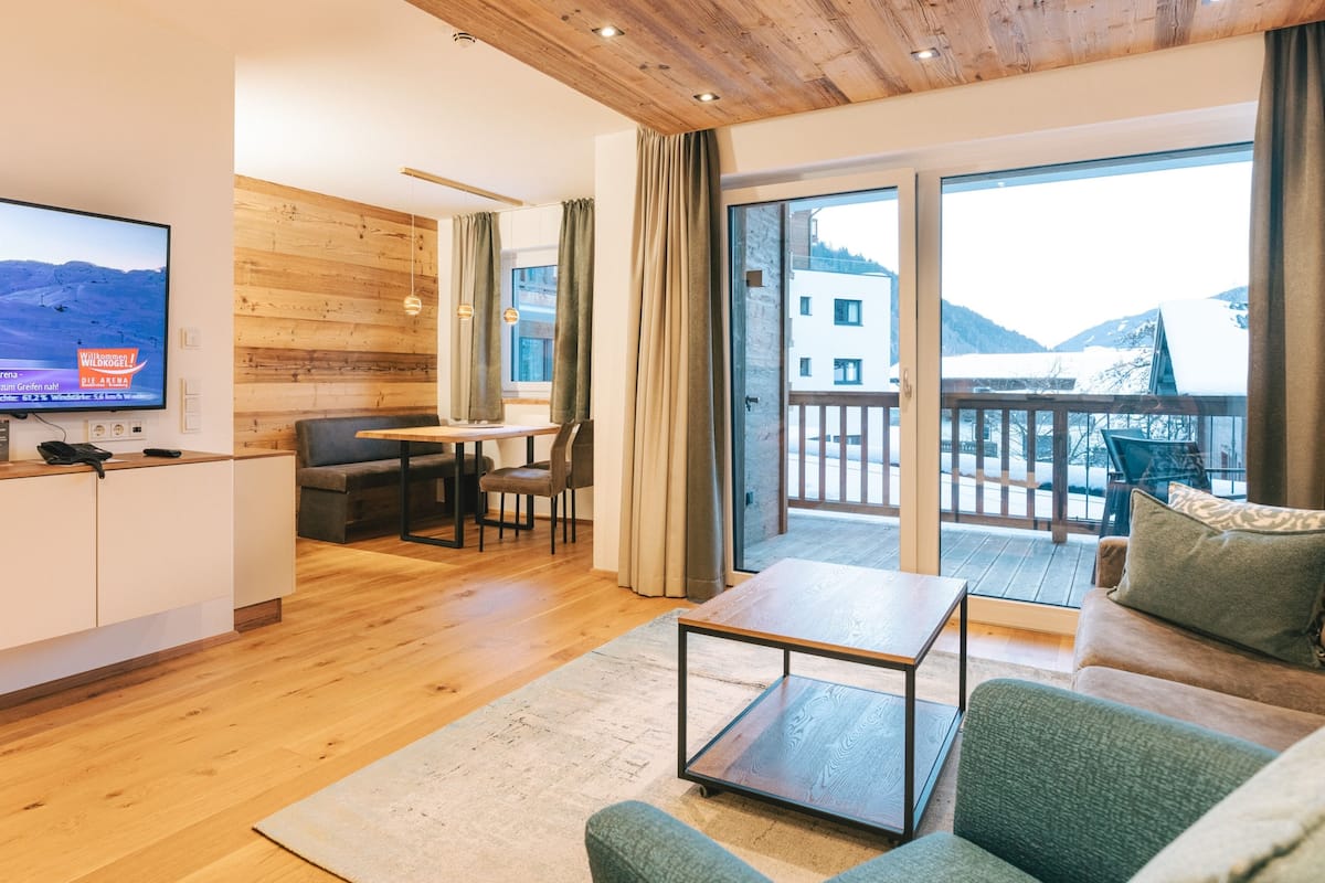 A modern living room with a wooden ceiling, a flat-screen TV, a sofa, a coffee table, and a balcony with a mountain view.