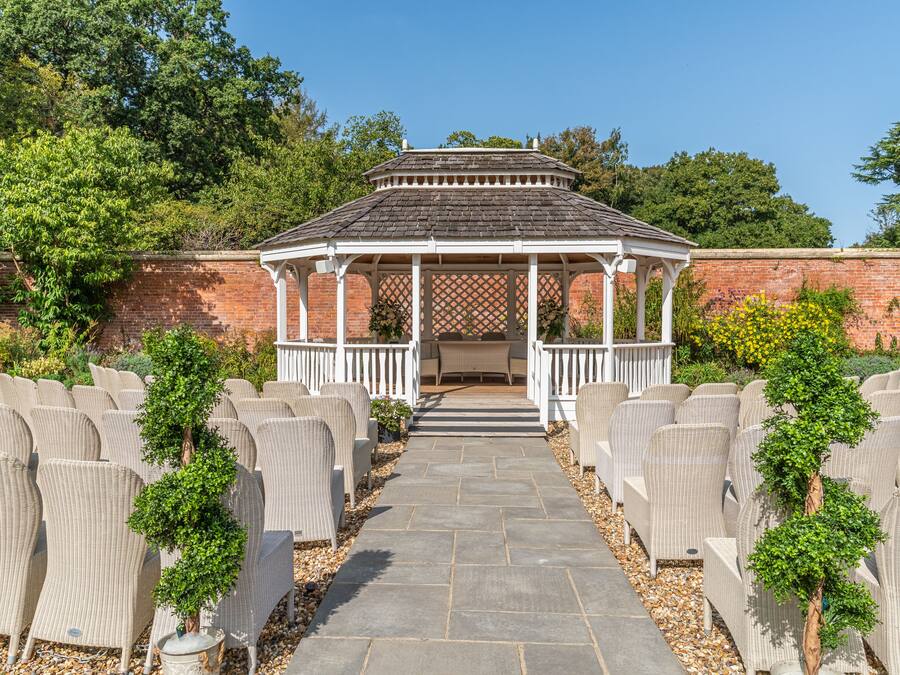 A gazebo with a tiled roof and white railings, surrounded by wicker chairs and small trees.