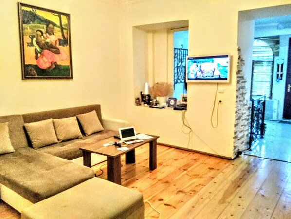A living room with a sectional sofa, a coffee table with a laptop, a TV mounted on the wall, and a wooden floor.