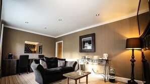 A modern hotel room with a dark-colored sofa, a small wooden coffee table, and a desk with a chair. There is a lamp on the right side and a framed picture on the wall. - Faenol Fawr Hotel (Rhyl)