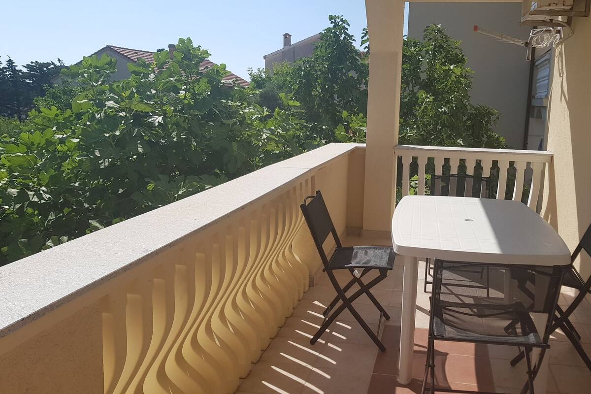 Un balcon avec une table blanche et deux chaises, une balustrade et une vue sur la verdure et les bâtiments.
