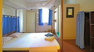 Un lit simple avec un matelas blanc et une couverture jaune, une tête de lit en bois, une petite table de chevet avec une lampe et une armoire avec un miroir.