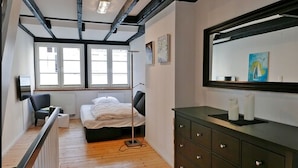 Ein Schlafzimmer mit Bett, Schminktisch und einem großen Spiegel. Fenster sorgen für natürliches Licht und unter den Fenstern befindet sich eine Heizung.