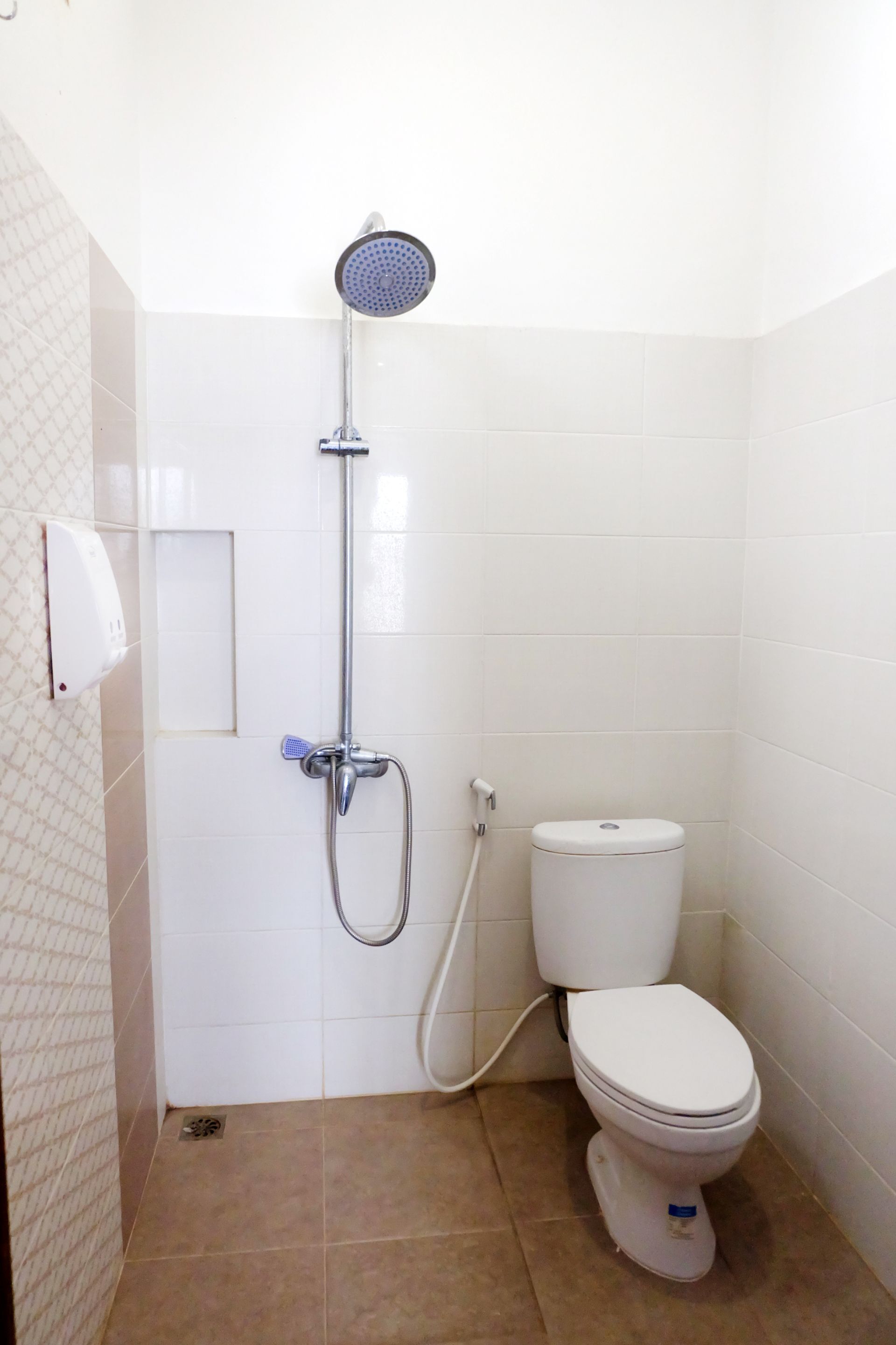 A small, clean bathroom with white tiles, a toilet, a showerhead, and a hand dryer.