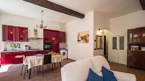 Un salon moderne avec un canapé blanc et des coussins bleus, un coin repas avec une table et des chaises, et une cuisine avec des meubles rouges et un lustre.