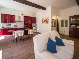 Una sala de estar moderna con un sofá blanco y cojines azules, un comedor con mesa y sillas, y una cocina con armarios rojos y una araña.