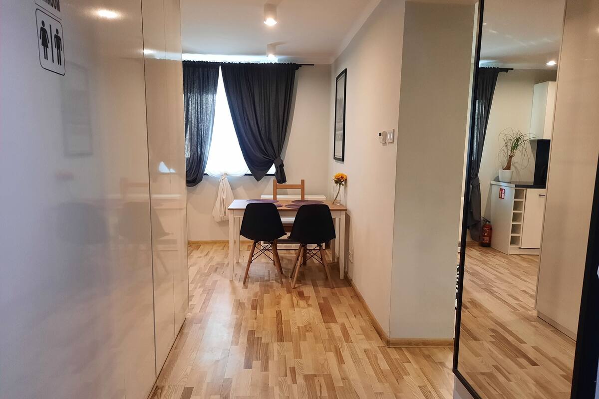 A modern, well-lit room with a wooden floor, a dining area with a table and chairs, and a visible bathroom sign.