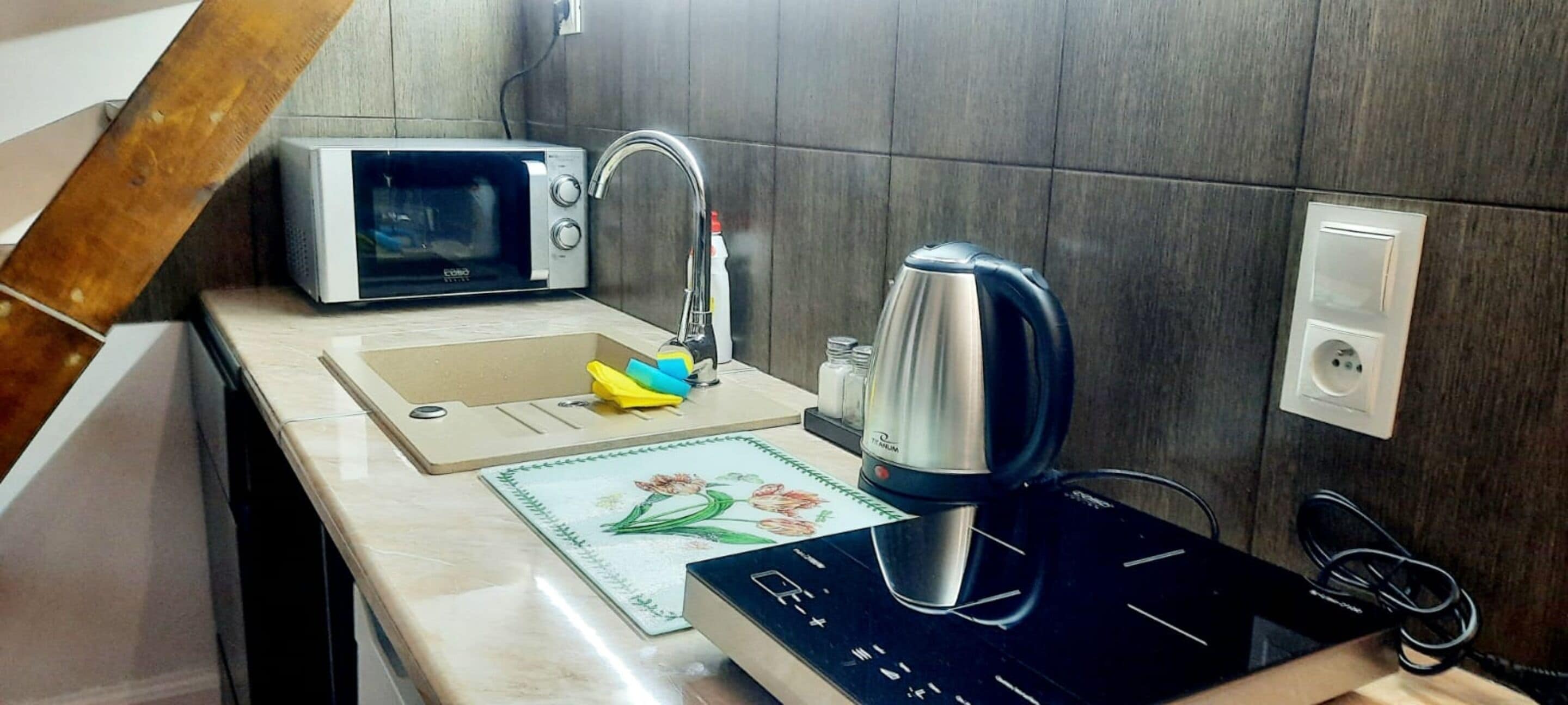 A kitchen with a microwave, kettle, and a sink with a cutting board and sponge.