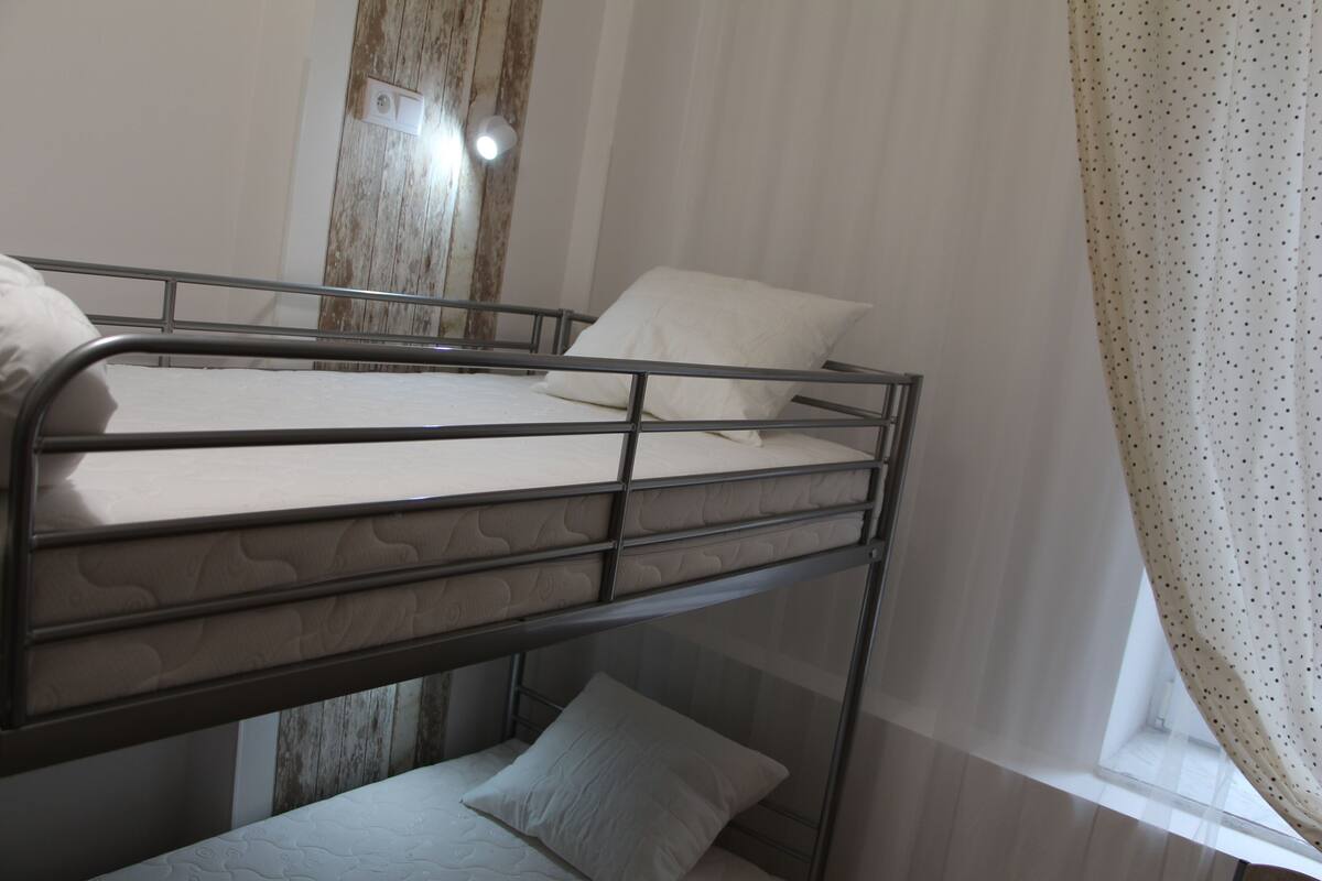 A bunk bed with a mattress and pillows, a wooden wall, and a window with curtains.