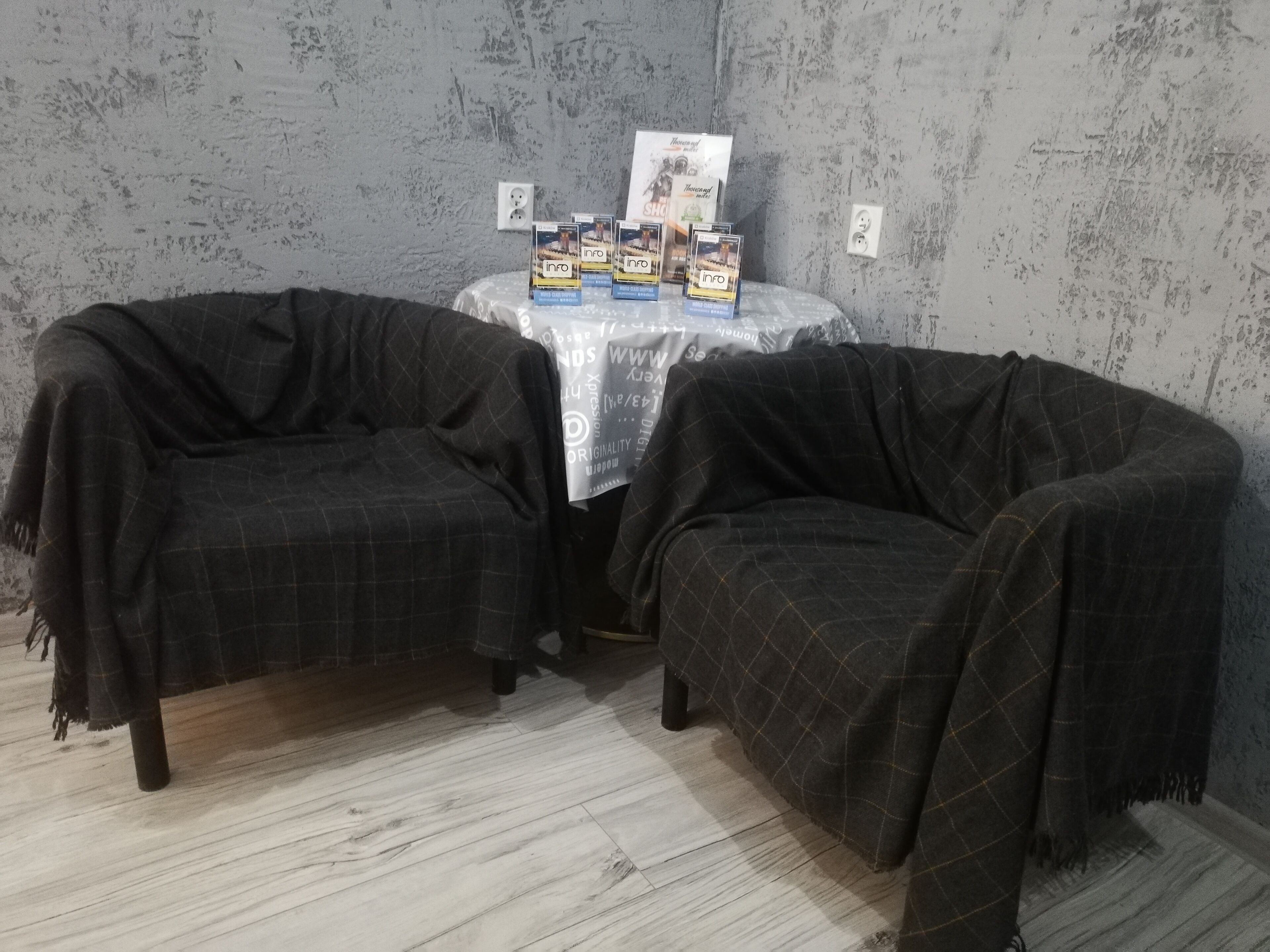 Una habitación con dos sillones tapizados en tela de cuadros oscuros, una mesita con folletos informativos y una pared gris con textura.