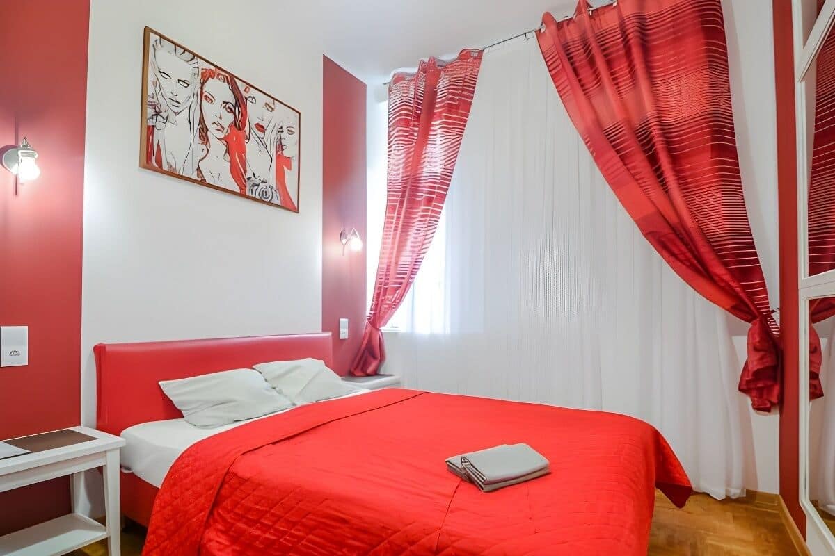 A bedroom with a red bedspread, white pillows, a red headboard, a nightstand, a red curtain, and a wall-mounted light fixture.
