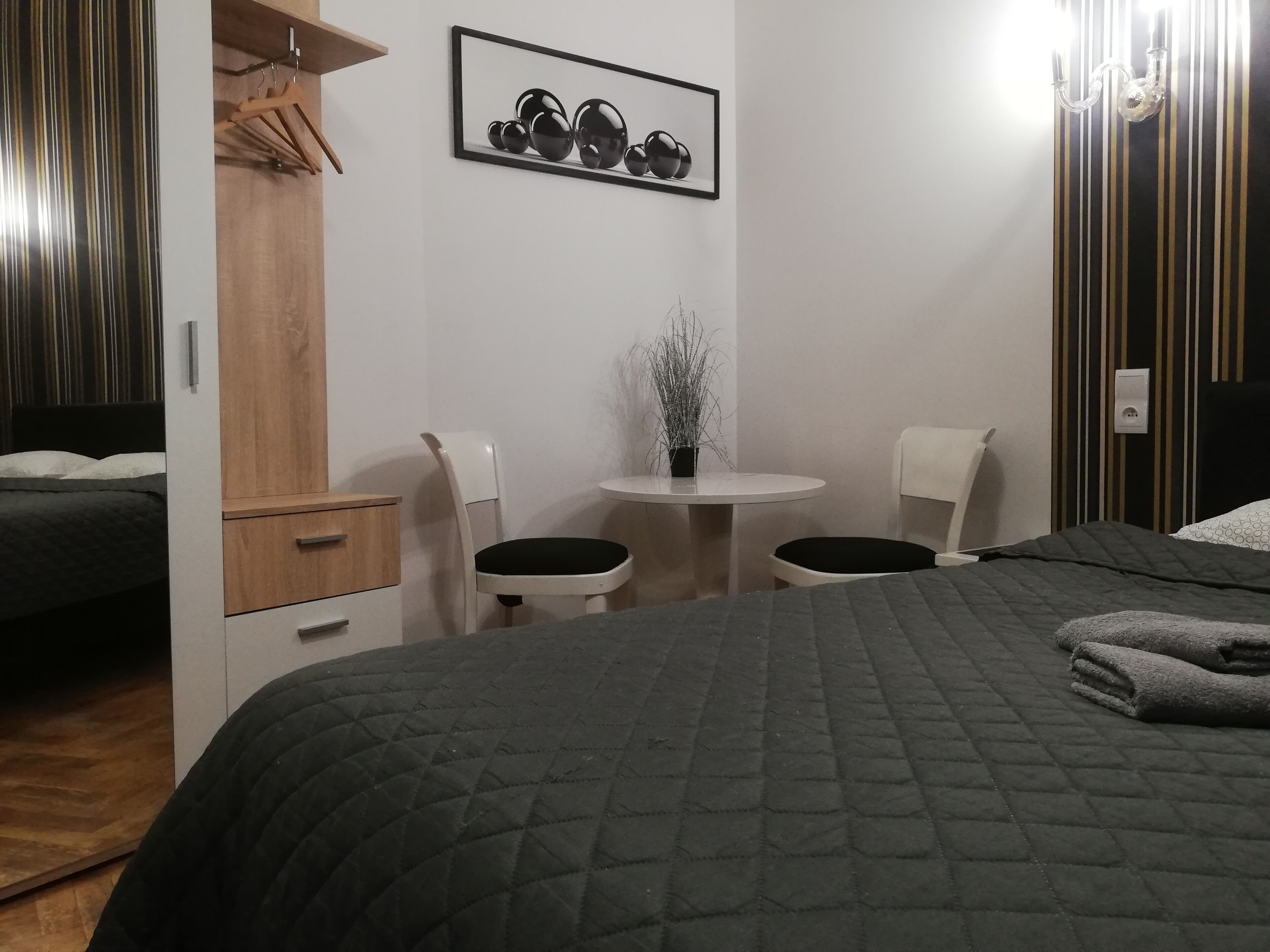 A bedroom with a bed, a desk, two chairs, a small table with a plant, and a wardrobe.
