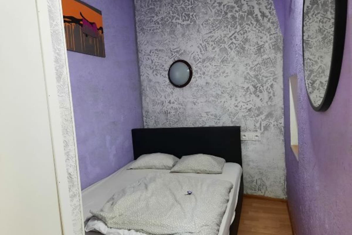 A small, narrow room with a bed, a round mirror, and a wall-mounted picture.