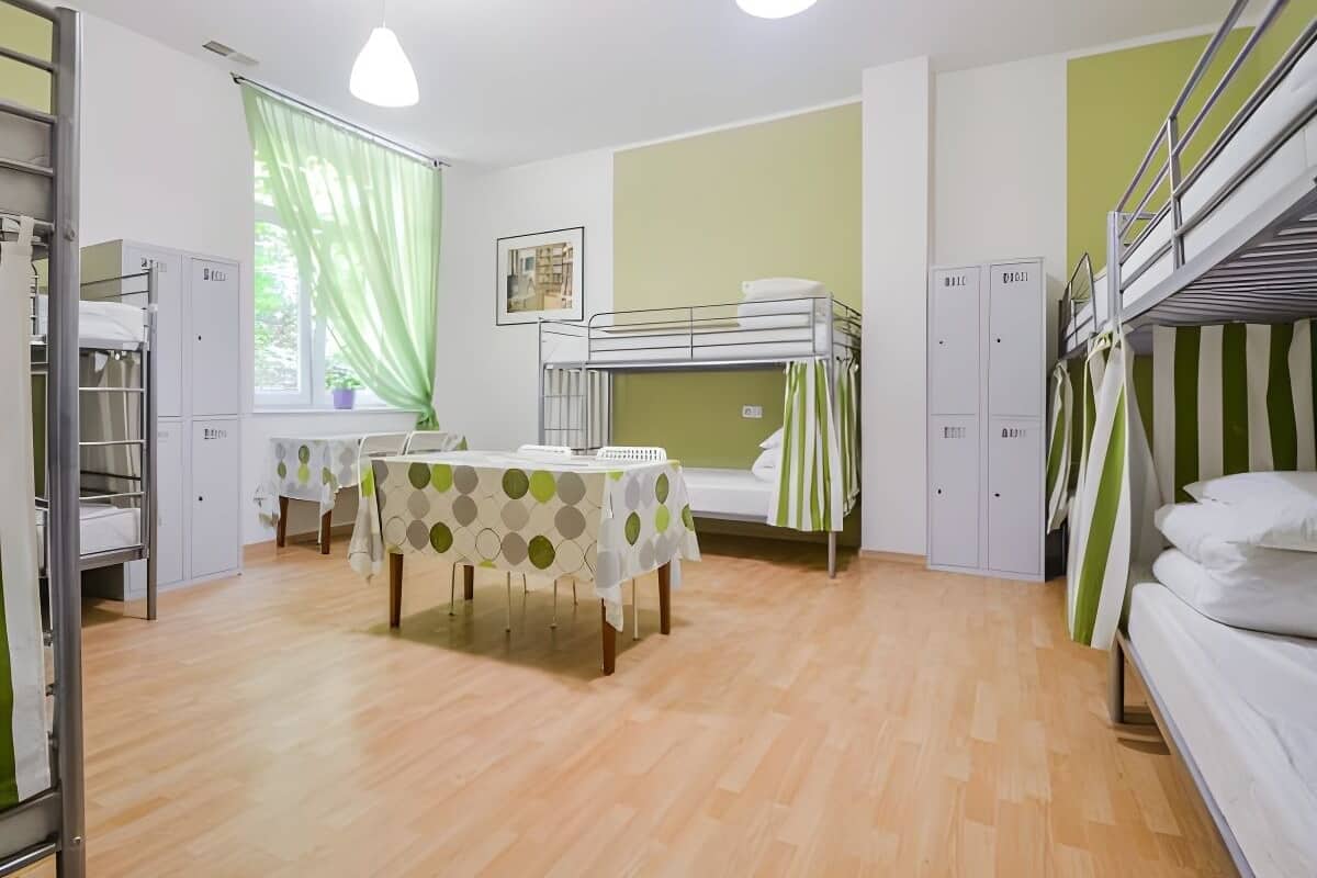 A room with bunk beds, a table with a patterned tablecloth, and a window with green curtains.