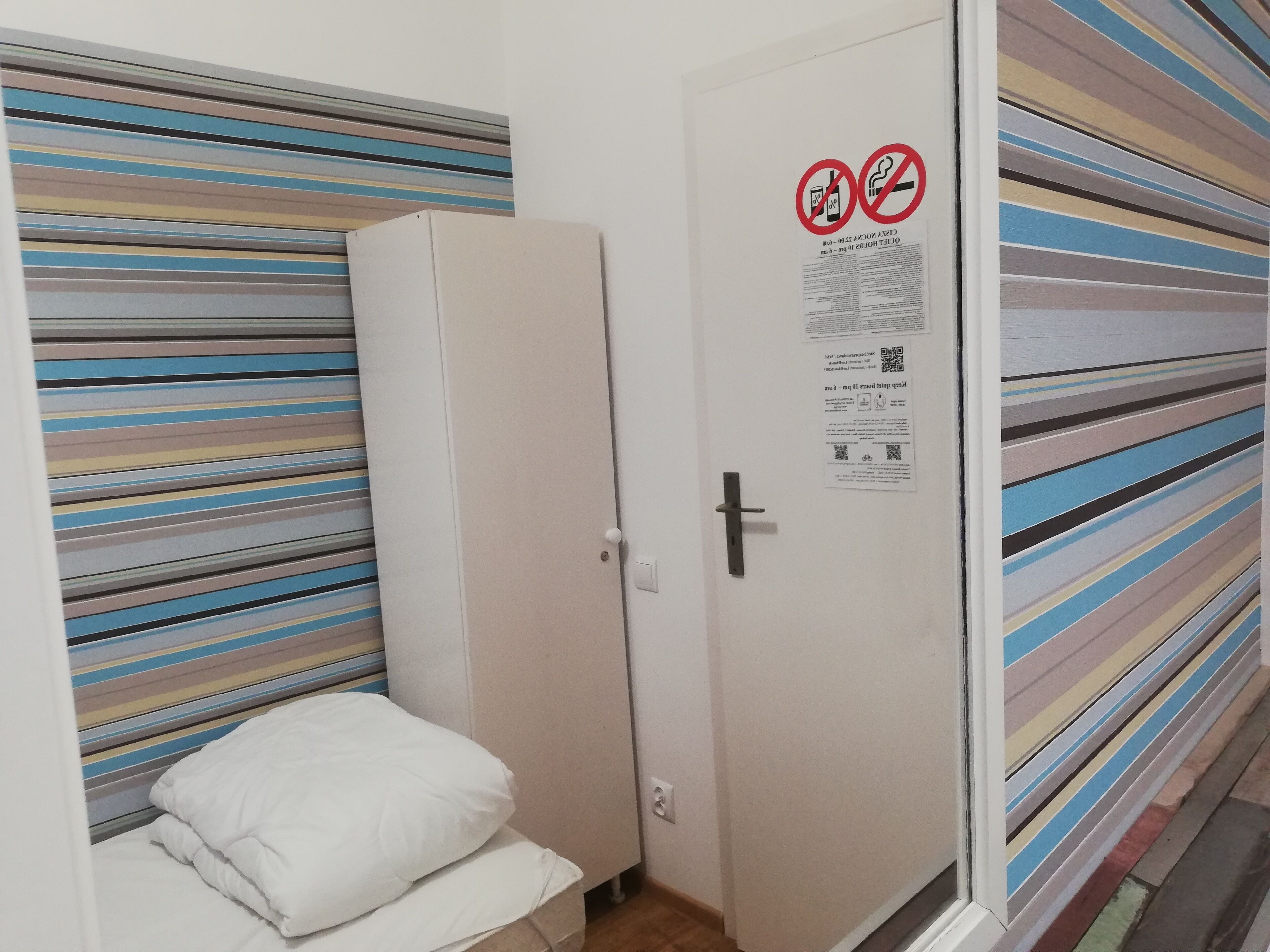 A hotel room with a striped headboard, a white door with a no-smoking sign, and a small bed with white bedding.