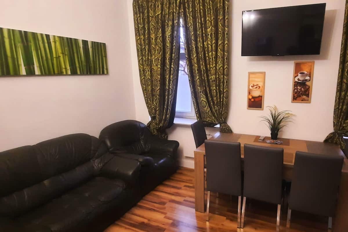 A living room with a black leather sofa, a wooden dining table with four chairs, a flat-screen TV mounted on the wall, and a window with patterned curtains.
