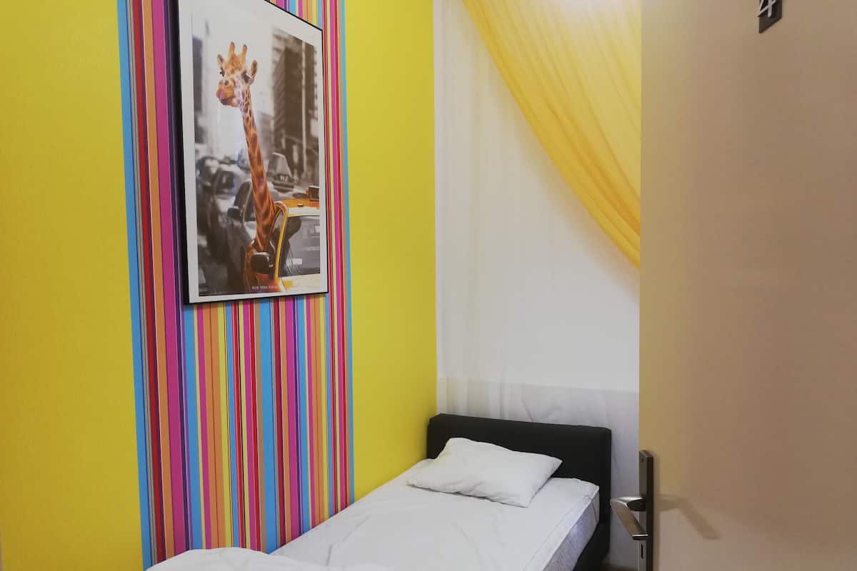 A small, single-bed room with a colorful striped wall, a framed picture of a giraffe, and a window with yellow curtains.
