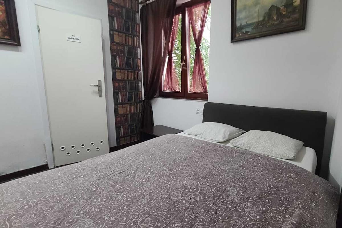 A bedroom with a bed, two pillows, a bookshelf, a door labeled "WC", and a window with curtains.