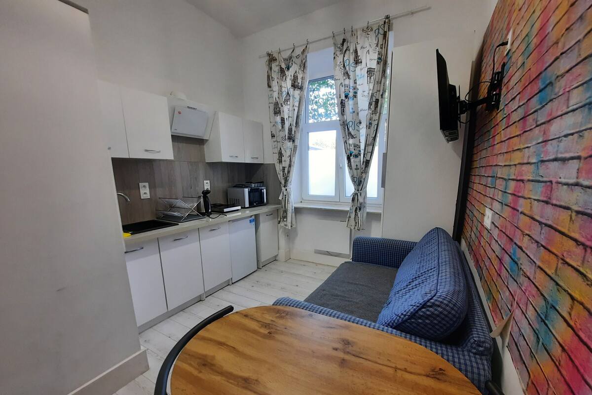 A compact kitchen with white cabinets, a microwave, and a sink. There's a wooden table and a blue sofa against a colorful brick wall.