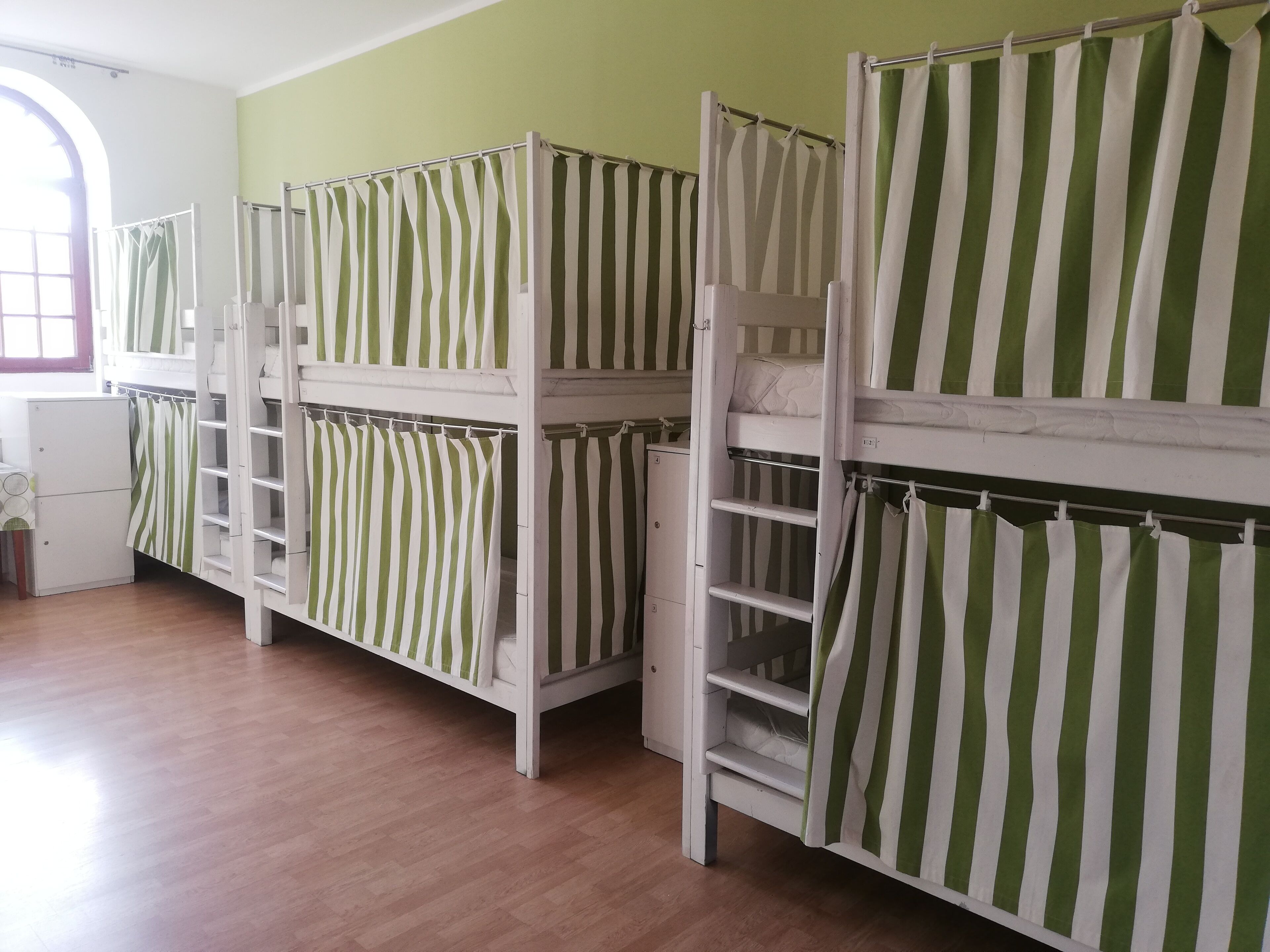 Camas literas con ropa de cama a rayas verdes y blancas, un armario blanco y suelo de madera en una habitación con ventana.